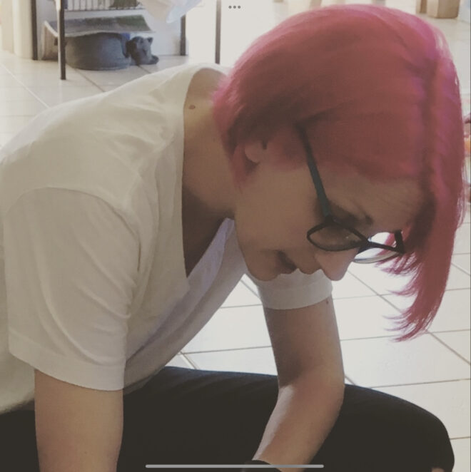 A female adult with hot pink hair and black rimmed glasses, sitting on the floor, leaning forwards.