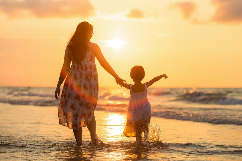 An adult in a dress holding the hand of a child in a dress, wading into the ocean in front of a low sun