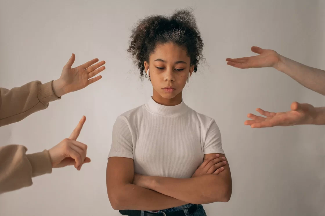 A girl with black hair, wearing a white t-shirt, surrounded by hands gesturing in critical ways