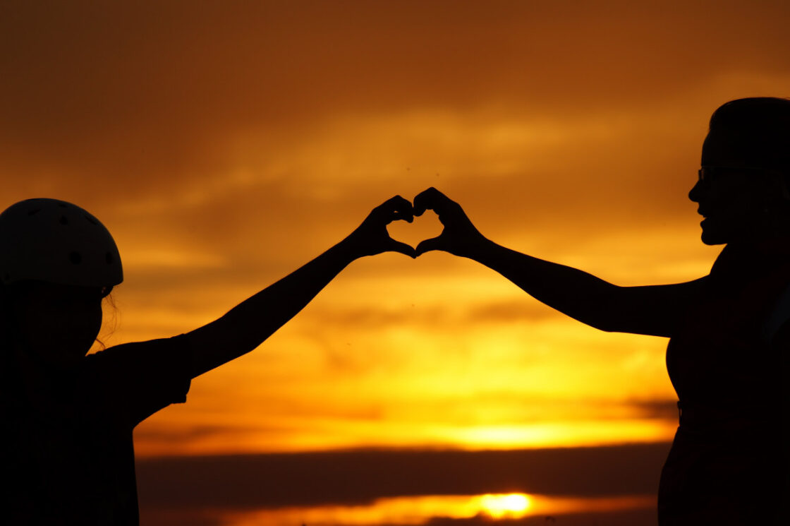 An adult and child reaching out their hands to form the shape of a heart, in front of a sunset