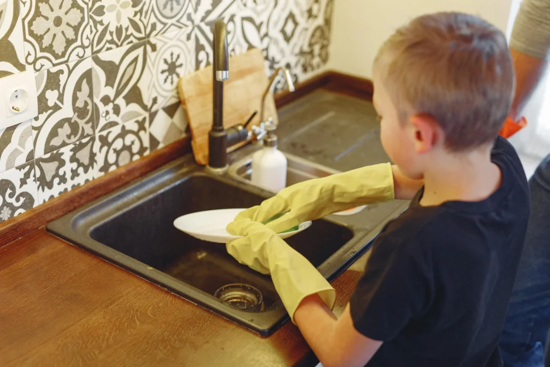 A boy wearing yellow gloves, washing dishes at a sink