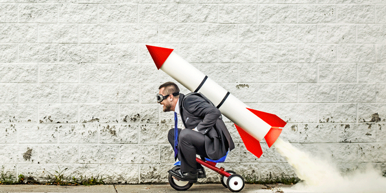 A man wearing goggles, on a small red tricycle, with a lit rocket tied to his back, demonstrating a lack of impulse control